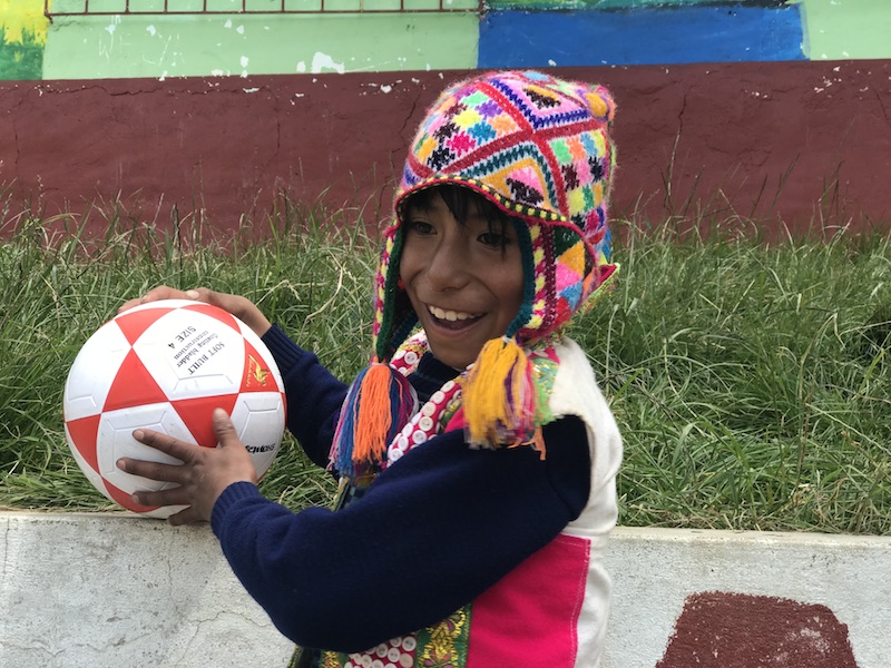 A small peruvian boy, wearing a colourful hat, smiles while holding a football