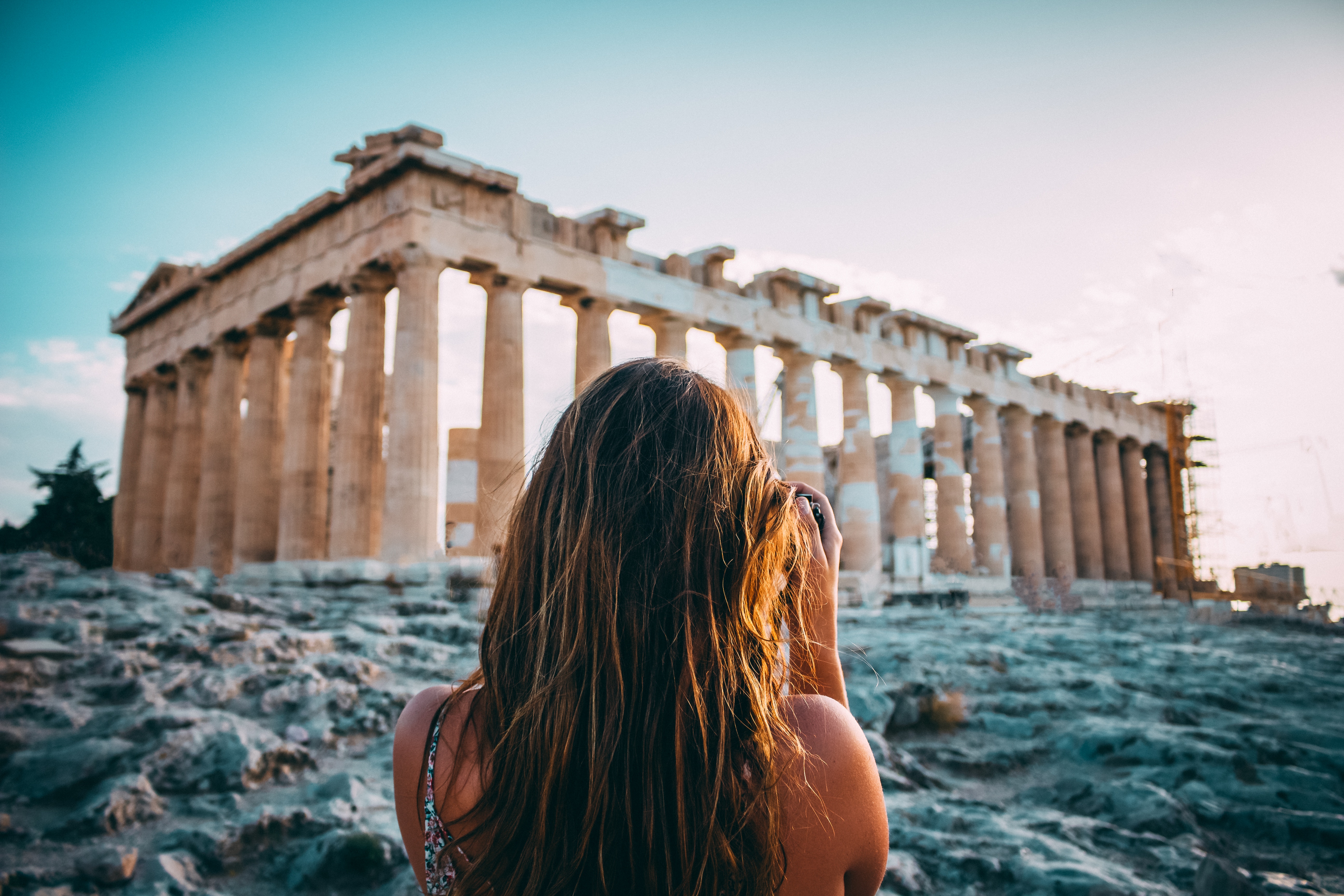 A woman with long hair, seen from behind, taking a photo of ruins in Greece
