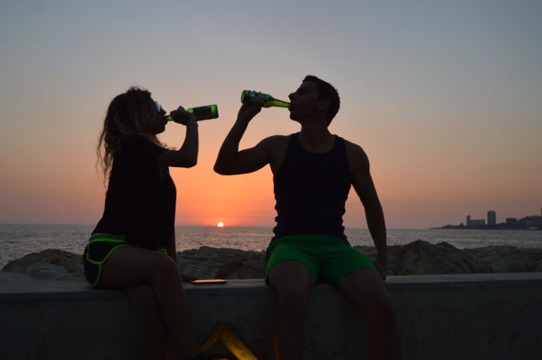 A man and a woman drinking beer in front of the sun setting above the ocean. 