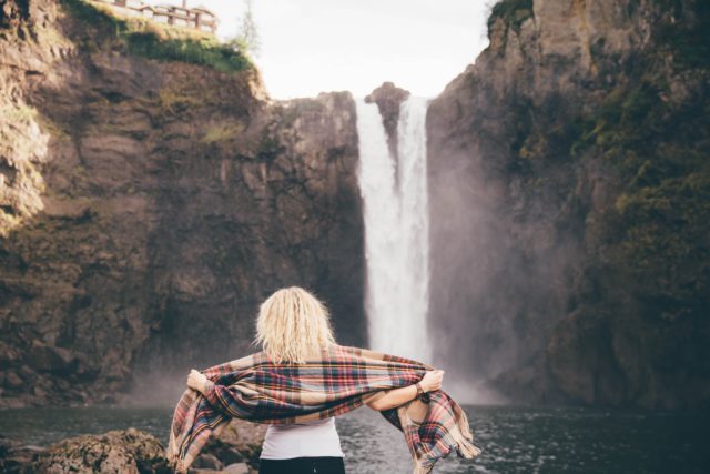 A blonde woman wraps her self in a scarf in front of a waterfall